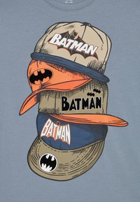 Grafiska T-krekls ar sakrautiem beisbola cepurēm krēmkrāsā, oranžā un jūras zilā, ar "Batman" logotipiem un sikspārņu motīviem dažādos stilus.
