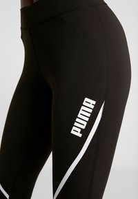 Black leggings made of stretchy fabric featuring a white logo on the side and a contrasting white stripe design along the seam.
