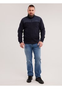 Man met baard, gekleed in een marineblauw jack met rits, lichtblauwe jeans en zwarte sneakers, staand tegen een effen lichte achtergrond.