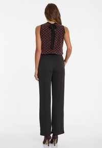 Zwarte jumpsuit met een gemusterde bordeaux rode top en een strikdetail op de rug, met wijde broekspijpen en hoge hakken met een textuurafwerking.