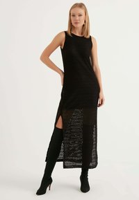 Abito midi senza maniche di colore nero con motivo a zigzag, sezione inferiore trasparente, spacco laterale, tessuto a maglia testurizzato, abbinato a stivali neri alti fino alla coscia.