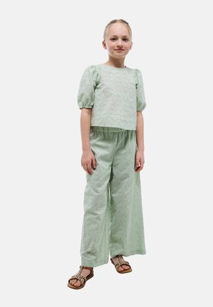 Jeune fille portant une tenue en tissu œillet vert clair avec des manches bouffantes et un pantalon large, debout avec une posture détendue et une expression neutre.