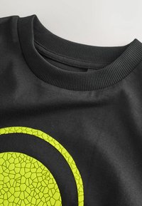 Fekete pulóver bordázott nyakkivágással és egy textúrált neon sárga kör alakú grafikával az elején, amely repedezett mintázattal rendelkezik.