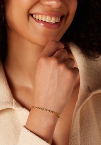 Gouden gedraaide armband om een pols met een lichte huidskleur, een close-up van een hand die op de kin rust, met textielachtige stof op de achtergrond.