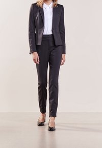 Costume noir taillé sur mesure avec une veste ajustée et un pantalon slim. La texture est lisse, assortie à un chemisier blanc à col et des talons vernis noirs.