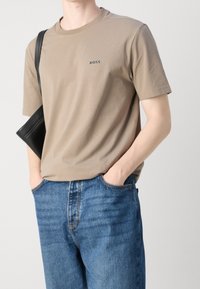 Světle hnědé bavlněné tričko s krátkými rukávy a kulatým výstřihem. Na hrudi má malé černé logo "BOSS" a je kombinováno s modrými džínami.