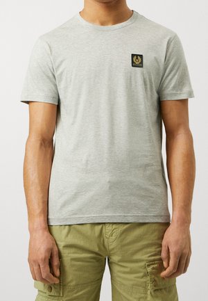Mand iført en lysegrå T-shirt med korte ærmer med lille sort og guld logo på brystet og olivengrønne cargoshorts.