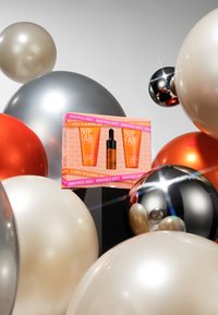 NIP + FAB Hautpflegeset in lebhaftem Packaging mit zwei orangen Tuben und einer dunklen Serumflasche, umgeben von metallischen und matten Ballons.