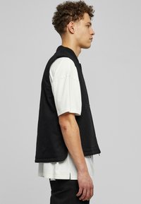 Melnas bezpaaka vests, kas izgatavots no teksturēta auduma, ar taisnu griezumu un divām priekšējām kabatām, uzvilkts virs baltas īsām piedurknēm kreklam.