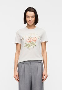 Jeune femme aux cheveux courts et foncés, portant un T-shirt blanc orné de pivoines roses et un pantalon gris plissé, debout devant un fond uni.