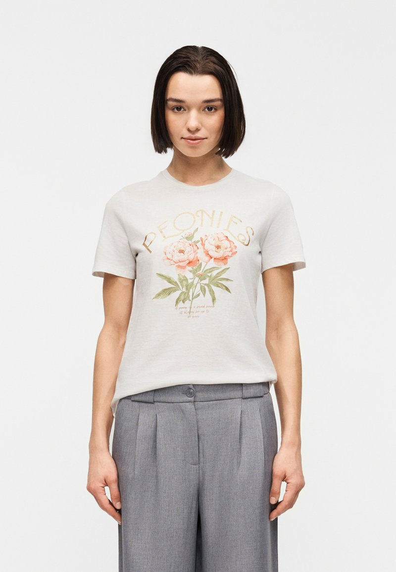 Jeune femme aux cheveux courts et foncés, portant un T-shirt blanc orné de pivoines roses et un pantalon gris plissé, debout devant un fond uni.