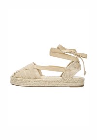 Beige pletená espadrilová sandála s kříženými pásky, šněrováním kolem kotníku a splétanou juteovou podrážkou. Otevřená špička s plochým gumovým podpatkem.
