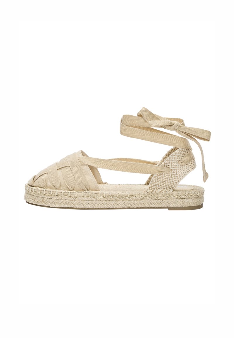 Beige pletená espadrilová sandála s kříženými pásky, šněrováním kolem kotníku a splétanou juteovou podrážkou. Otevřená špička s plochým gumovým podpatkem.