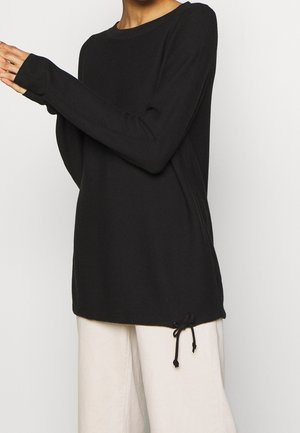 Persoon draagt een zwart trui met lange mouwen en een zijstrik, gecombineerd met losse witte broek.