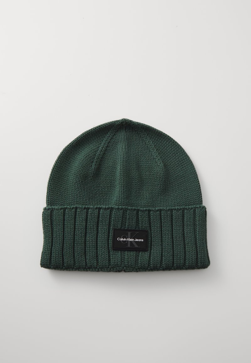 Gorro de malha verde com punho canelado, apresentando uma etiqueta preta com texto branco "Calvin Klein Jeans." Textura suave, design de topo arredondado.