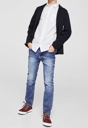 Blaue Jeans aus Denim, weißes Hemd mit Knöpfen, schwarzer Blazer und bordeauxrote Sneaker mit weißen Sohlen. Lässiges, geschichtetes Outfit mit kontrastierenden Farben.