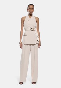 Top beige con scollo a halter e cintura con fiocco, abbinato a pantaloni a gamba larga nello stesso tessuto. Texture liscia e design minimal.