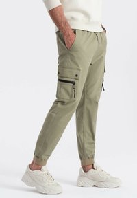 Osoba w beżowych spodniach cargo z bocznymi kieszeniami i kremowych sneakersach, stojąca z rękami w kieszeniach na białym tle.