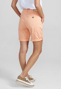 Pantalones cortos enrollados en color durazno con bolsillos traseros y detalles de botones. Hechos de una tela ligera, con una textura sutil y acentos de costura.