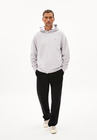 Sweatshirt à capuche gris clair avec une poche avant, porté sur une chemise blanche ; associé à un pantalon noir et des baskets crème. Design minimal.