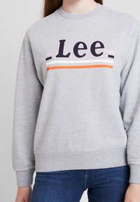 Sudadera gris con cuello redondo, que presenta 'Lee' en letras negras en negrita y rayas coloridas en rosa, blanco y naranja a lo largo del pecho.