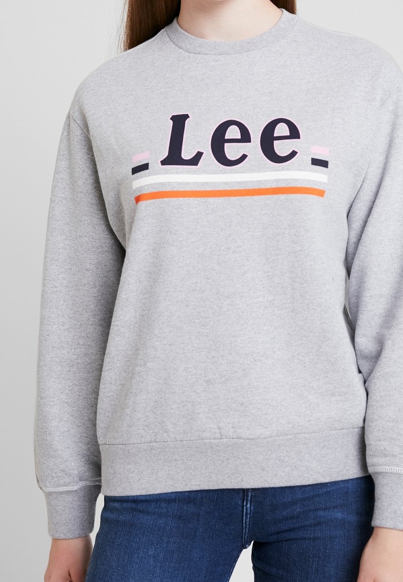 Sudadera gris con cuello redondo, que presenta 'Lee' en letras negras en negrita y rayas coloridas en rosa, blanco y naranja a lo largo del pecho.