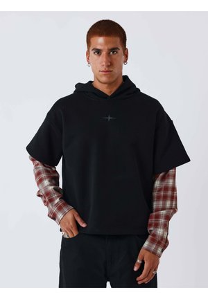 Joven con cabello rojo corto que lleva pendientes de aro, una camiseta negra con capucha de manga corta sobre una camisa de cuadros roja de manga larga y anillos plateados.