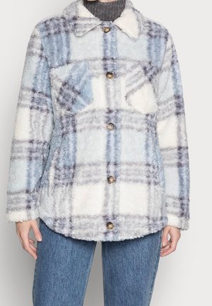 Blød, fluffy ternet jakke i hvid, blå og grå med knapper foran og brystlommer, båret over en mørk rullekrave og blå jeans.