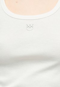 Ασημένιο κολιέ με δύο μενταγιόν σε σχήμα πουλιού, φορεμένο πάνω από λευκό ριμπαντινό τοπ σε γυμνό δέρμα.