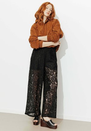 Femme aux cheveux roux appuyée contre un mur blanc, portant une veste en daim marron, un pantalon large en dentelle noire et des sandales à plateforme noires.