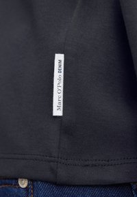 Mørkegrå T-shirt med et hvidt mærke på sidesømmen, blød tekstur, med synlige sømme og denimstof nedenunder.