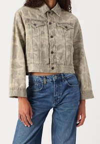 Afskåret olivengrøn denimjakke med et subtilt blomstermotiv, knaplukning og to brystlommer. Matchet med højtaljede blå jeans.