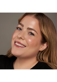 Primer plano de una persona con cabello largo y ondulado de color castaño claro, piel clara y maquillaje natural, sonriendo contra un fondo neutro.