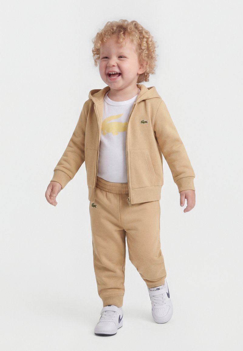 Niño pequeño con cabello rizado sonriendo, vistiendo una sudadera con capucha y cremallera color beige, pantalones a juego, camiseta blanca con logo amarillo y zapatillas blancas, caminando hacia adelante.