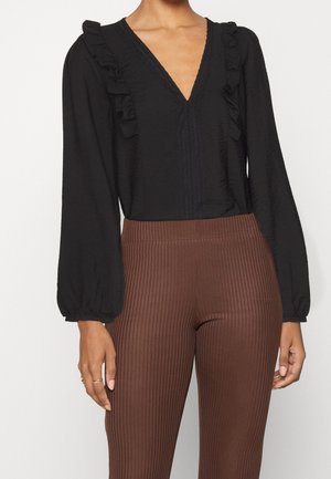 Vrouw die een zwarte blouse met lange mouwen en ruches draagt, gecombineerd met een bruine ribgebreide hoge taillebroek, staand tegen een witte achtergrond.