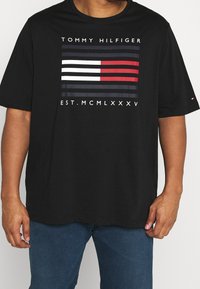 Muž v černém tričku Tommy Hilfiger s logem na hrudi, které je tvořeno námořnickými, bílými a červenými pruhy, a římskými číslicemi "EST. MCMLXXXV" nad modrými džíny.