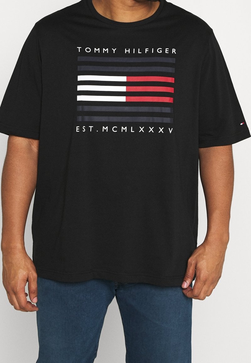 Muž v černém tričku Tommy Hilfiger s logem na hrudi, které je tvořeno námořnickými, bílými a červenými pruhy, a římskými číslicemi "EST. MCMLXXXV" nad modrými džíny.
