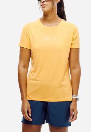 Femme portant des lunettes de soleil, un t-shirt de sport jaune à manches courtes, un short bleu marine et une montre blanche au poignet, debout devant un fond clair uni.