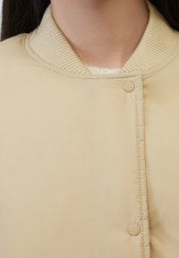 Giacca imbottita beige con colletto a coste, dotata di chiusura con bottoni automatici e texture liscia, che mette in evidenza un design minimalista.