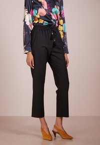 Pantalons noirs coupés avec une taille élastique et un cordon de serrage, assortis à un blouse à manches longues à motif floral et des chaussures à talons couleur moutarde.