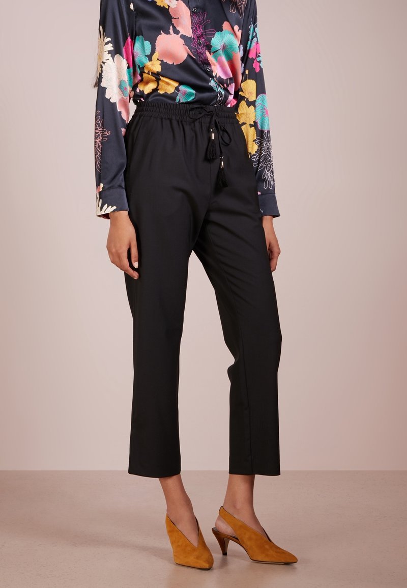 Pantalons noirs coupés avec une taille élastique et un cordon de serrage, assortis à un blouse à manches longues à motif floral et des chaussures à talons couleur moutarde.
