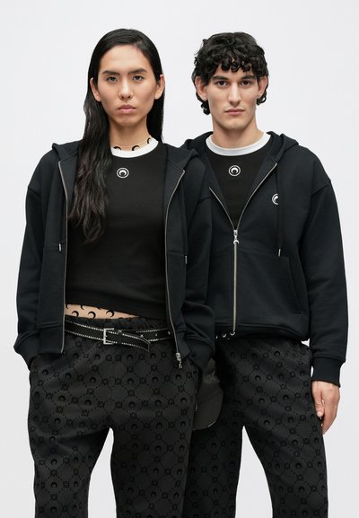 Sweatshirt preto com fecho de correr, camisa preta ajustada e calças com padrão circular. Apresenta tecido texturizado e detalhes em contraste.