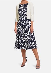 Blommönstrad klänning i marinblått och vitt, med en utsvängd kjol, rund halsringning och en ljus krämfärgad kofta med trekvartslånga ärmar.