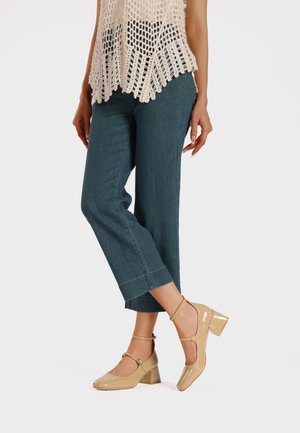 Décolleté in pelle verniciata beige con cinturino alla caviglia, punta quadrata e tacco spesso, abbinati a pantaloni cropped in denim blu. Top all'uncinetto con lavorazione aperta.