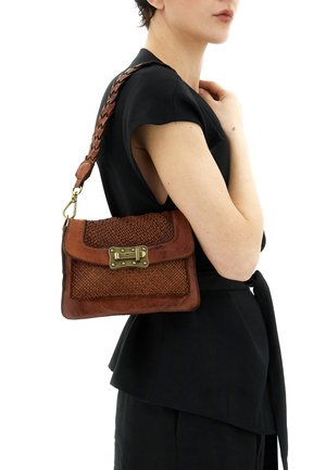 Donna che indossa un abito nero porta una borsa a tracolla in pelle marrone intrecciata con chiusura dorata sulla spalla destra.