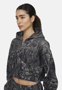 Felpa corta in grigio scuro con un motivo di camuffamento di rami e foglie. Ha un cappuccio con cordini e una consistenza morbida.