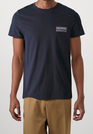 Mann trägt marineblaues T-Shirt mit kurzen Ärmeln und dem "Hilfiger Established 1985 USA"-Logo auf der linken Brust sowie beigefarbene Hose, steht vor hellem Hintergrund.