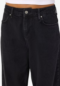 Crne traperice visokog struka s pet džepova, metalnim gumbom i suptilnim izblijedjenjem. Srednji struk s klasičnim ravnim krojem nogavica.