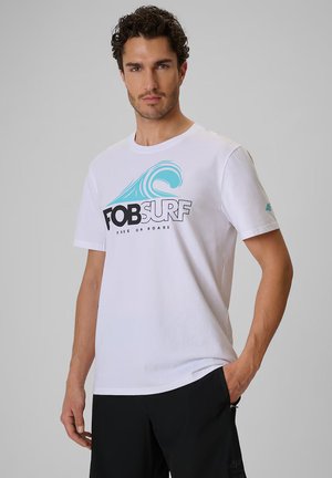 Man draagt een wit T-shirt met blauwe golf en zwarte tekst "FOB SURF" staande tegen een effen grijze achtergrond.