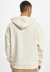 White hooded sweatshirt with a relaxed fit, ribbed cuffs, and hem. The fabric appears soft with a smooth texture. Lacks visible graphics.